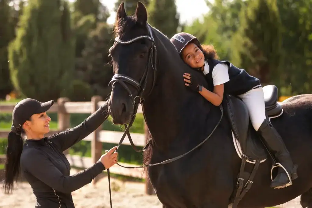 Bambina con cap abbraccia il collo di un cavallo morello mentre l'istruttrice lo tiene per le redini nel campo esterno