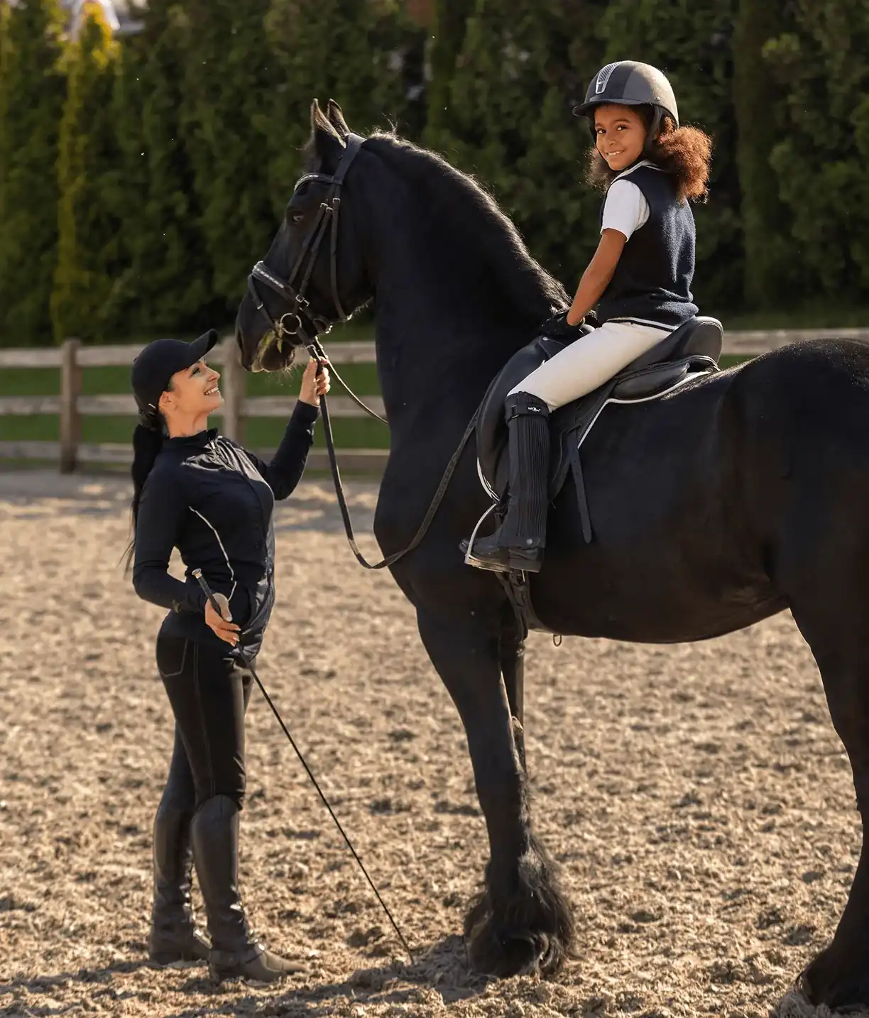 Istruttrice guida una bambina sorridente in sella a un cavallo nero nel campo