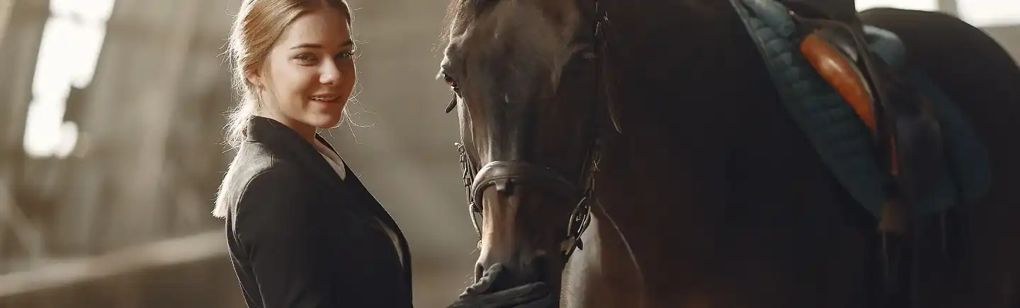Allieva sorridente accanto al suo cavallo baio sellato prima della lezione di equitazione in scuderia