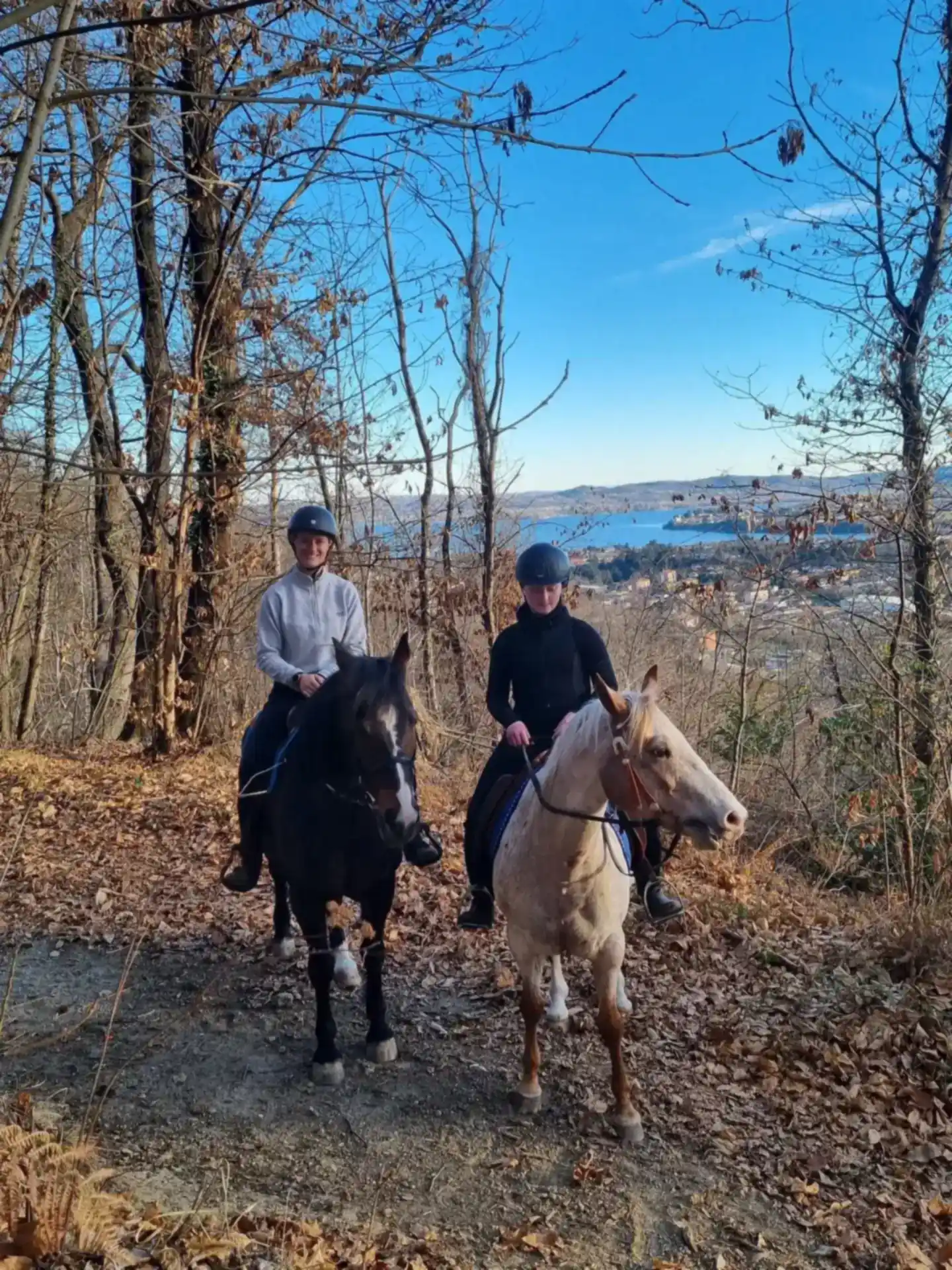 Due amazzoni sorridenti a cavallo su un sentiero nel bosco con il lago sullo sfondo