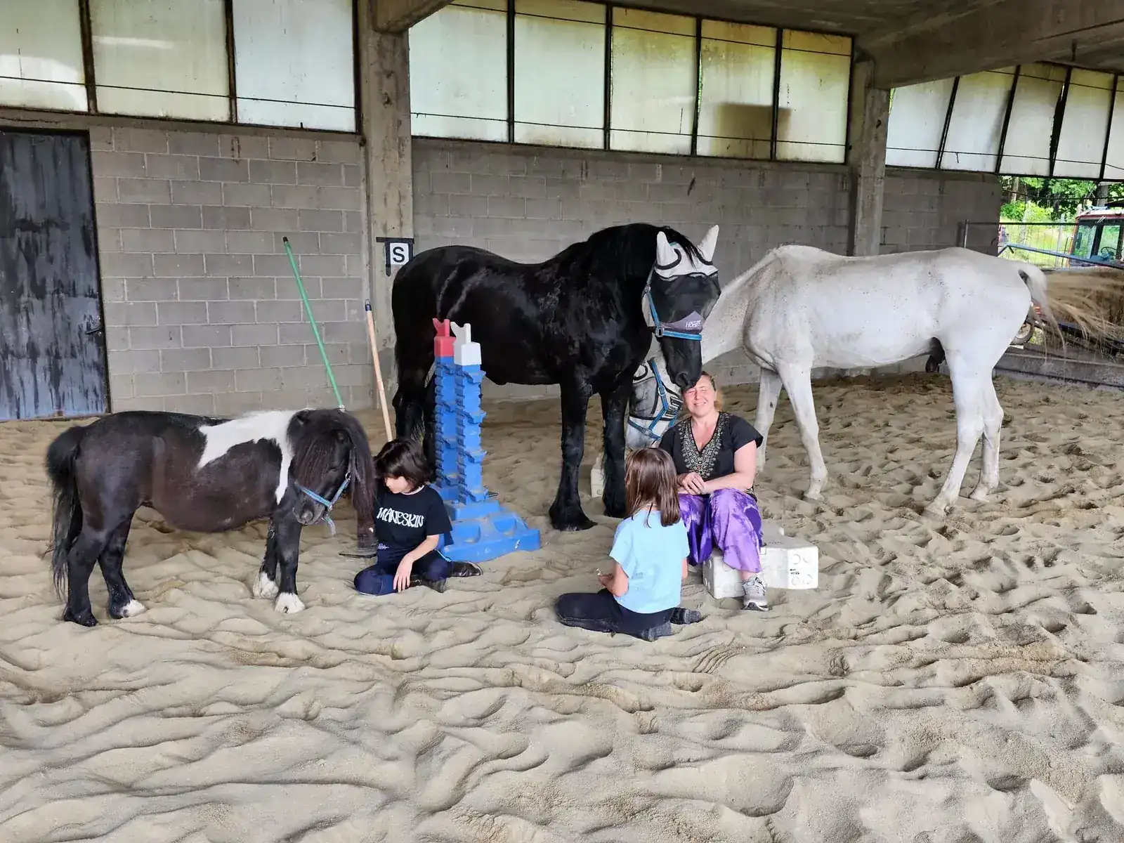 Due bambini seduti sulla sabbia del maneggio coperto insieme a pony, cavalli e istruttrice