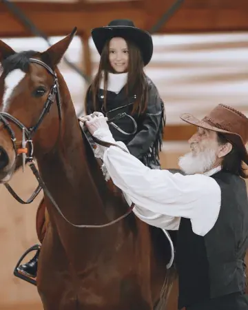 Bambina con cappello western sorride in sella a un cavallo baio mentre l'istruttore le sistema le redini nel campo coperto