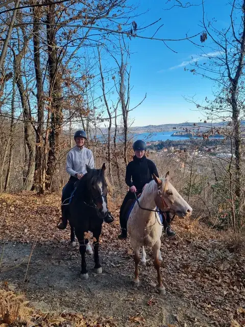 Due amazzoni sorridenti a cavallo su un sentiero nel bosco con il lago sullo sfondo