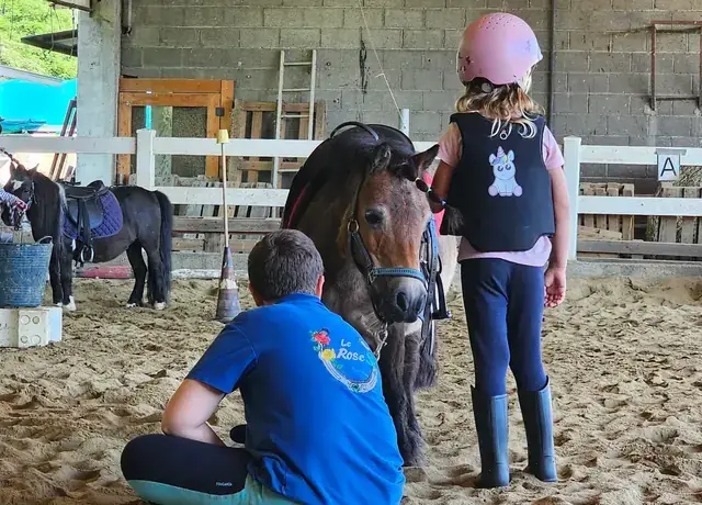 Bambina con casco rosa accanto al pony mentre un istruttore in polo Mitena Ranch la affianca