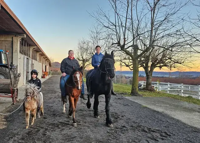 Bambino conduce un pony mentre due amazzoni escono a cavallo dalle scuderie al tramonto